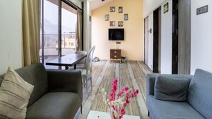 A modern living room with a sofa, a coffee table with remote controls, a TV on the wall, and a dining area with a table and chairs.
