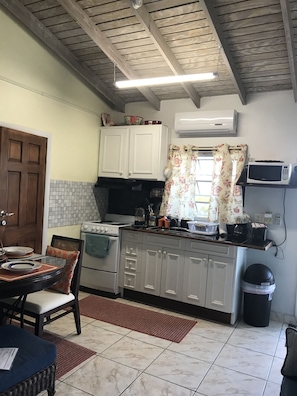 Compact kitchen with stove, microwave, and countertop area, featuring a window with floral curtains and a chair with cushion.