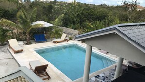 Una piscina con sillas de descanso y un área de estar cubierta, rodeada de follaje.