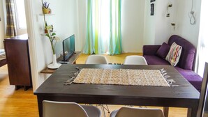 Un comedor con una mesa de madera oscura, sillas blancas, un mantel tejido y un sofá morado con un cojín colorido.