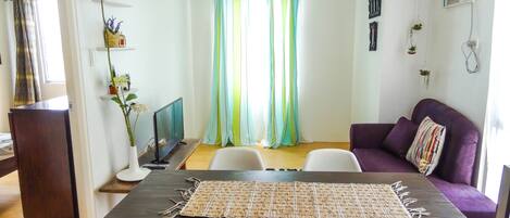 Un comedor con una mesa de madera oscura, sillas blancas, un mantel tejido y un sofá morado con un cojín colorido.