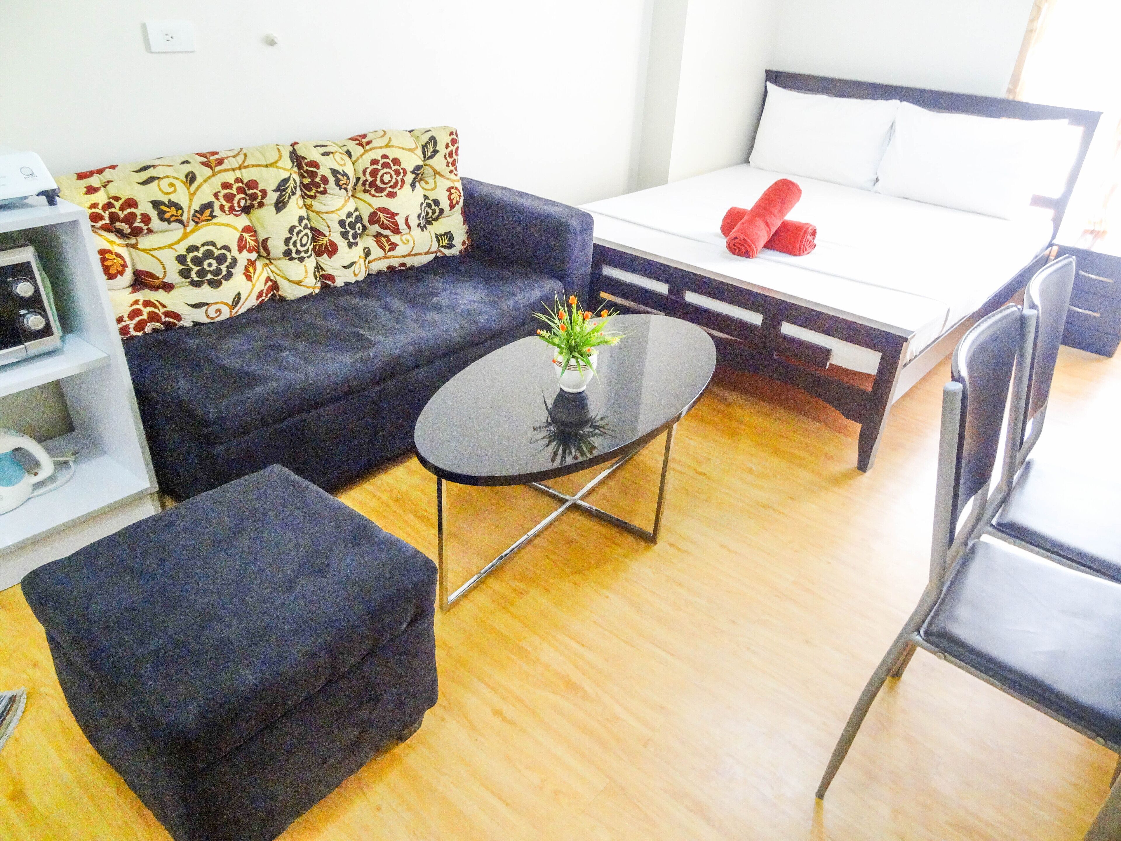 A modern room with a sofa, a small table with a plant, a bed with rolled towels, and a chair.