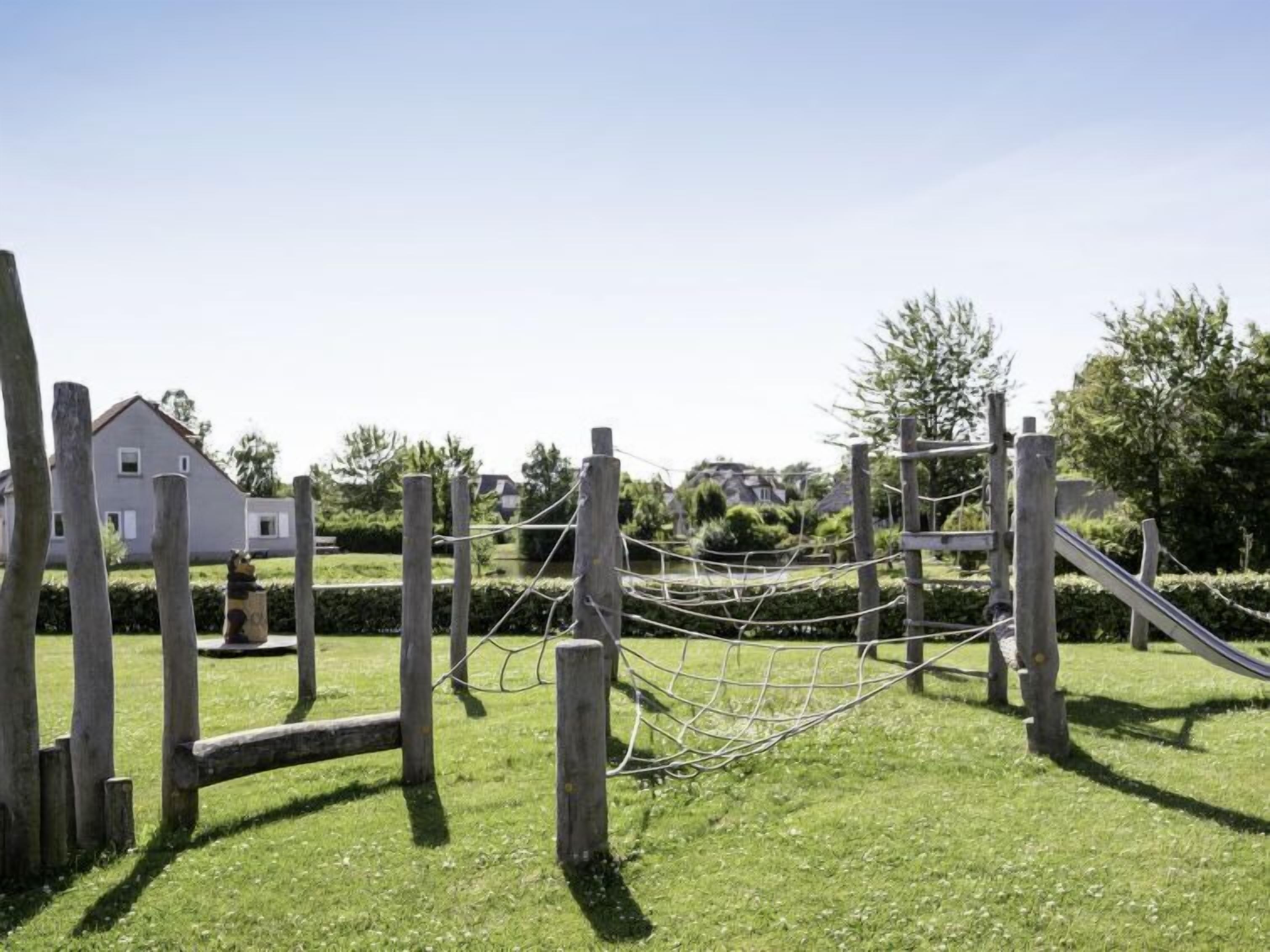 Une aire de jeux avec un pont de corde et un toboggan, entourée d’une clôture en bois et d’une pelouse.