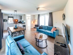 A modern living room with a sofa, a coffee table, and a TV. There is a dining area with a table and chairs. The room has a kitchen in the background.