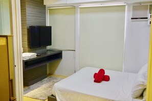 Una habitación de hotel con cama, televisor montado en la pared, escritorio con silla y estante con toallas dobladas.