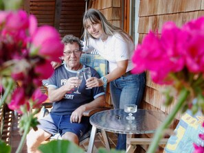 Una pareja de edad avanzada sentada en una pequeña mesa redonda con una copa de vino, en un entorno al aire libre de madera con un muro de madera al fondo.
