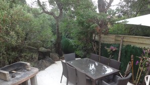 Een terras met een tafel met glazen blad en rieten stoelen, omgeven door groen en een stenen barbecue.