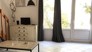 Een kamer met een bed, een tafel met een glas en een kaars, een televisie op een kast, en een raam met gordijnen.