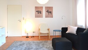 Un salon moderne avec un plancher en bois, un canapé noir, une chaise blanche et un tapis gris. On y trouve deux œuvres d’art murales représentant des élans, une lampe sur pied et un luminaire au plafond.