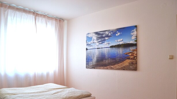 Une chambre à coucher avec un lit, une fenêtre avec des rideaux et un tableau représentant un paysage lacustre accroché au mur.