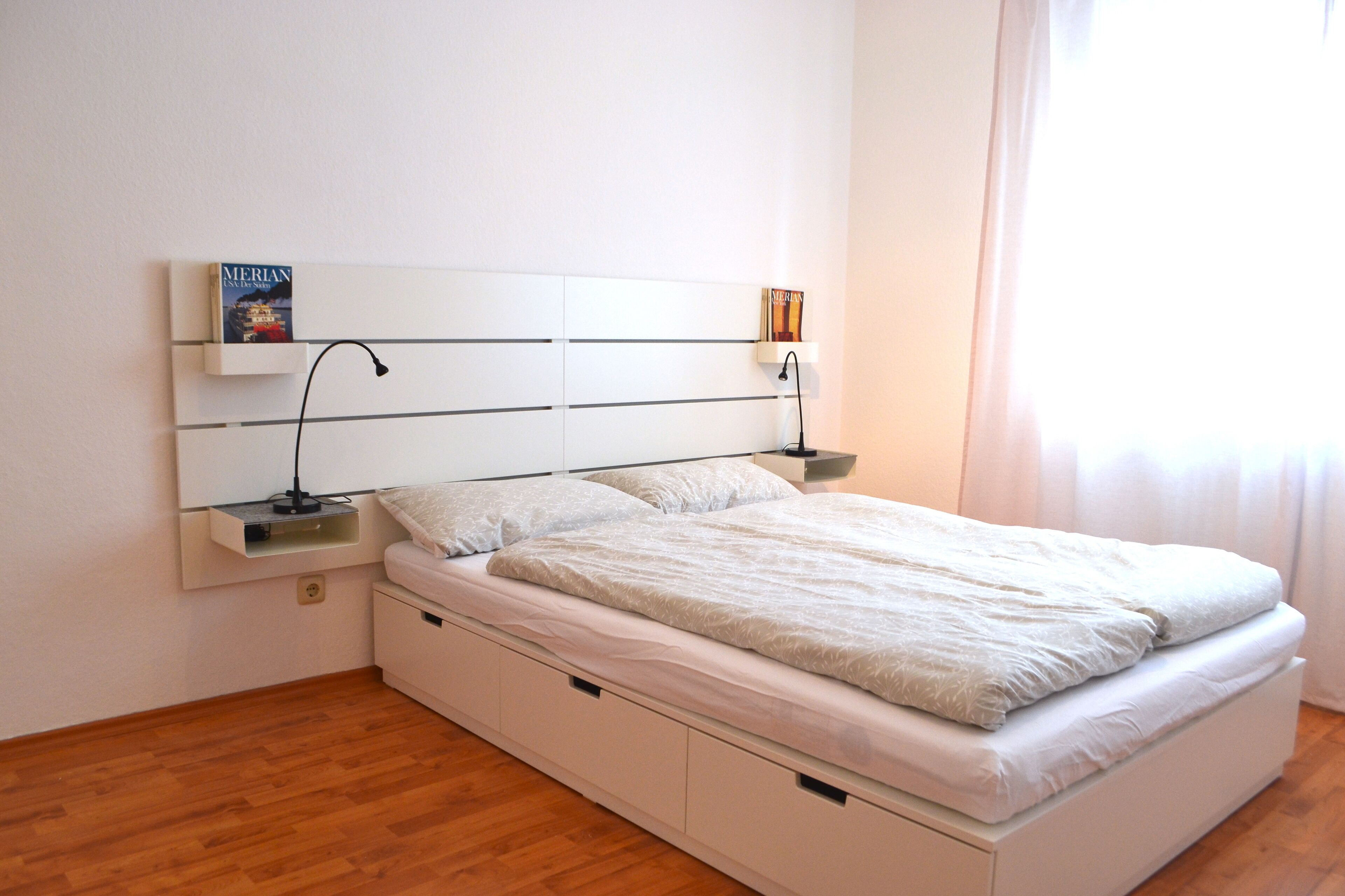 A modern bedroom with a white bed, built-in nightstands, and a wall-mounted shelf with books.