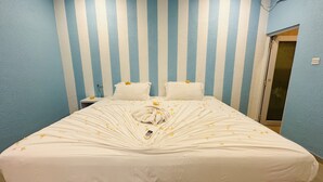 A hotel room with a striped blue and white wall, a neatly made bed with a white comforter and pillows, and a small bedside table with a bottle and a glass. - Midsummer Thulusdhoo (Thulusdhoo Island)