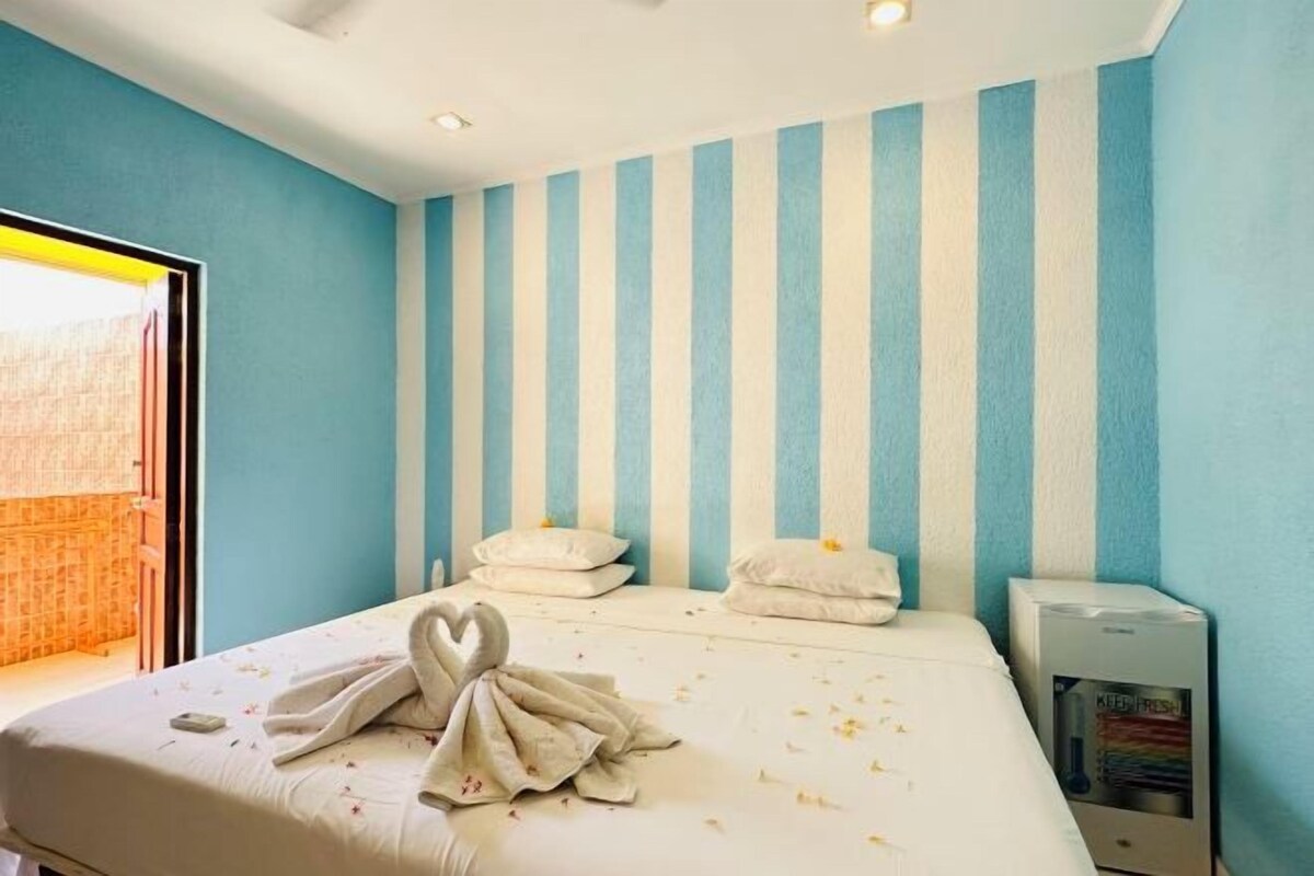 A hotel room with a striped wall, a bed with a heart-shaped towel, and a minibar.