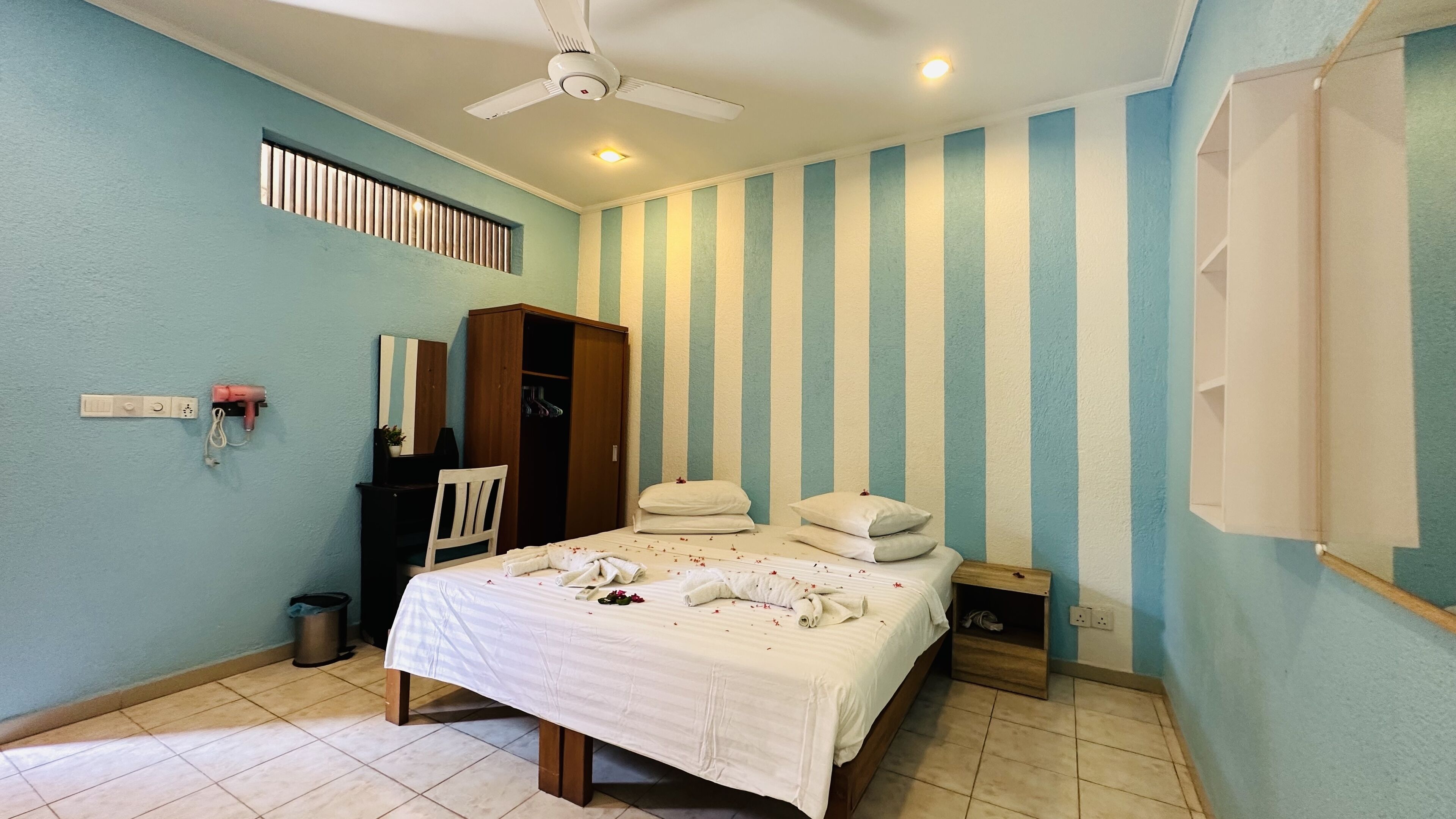 A bedroom with a bed, a wardrobe, a ceiling fan, a window with blinds, and a wall-mounted shelf.