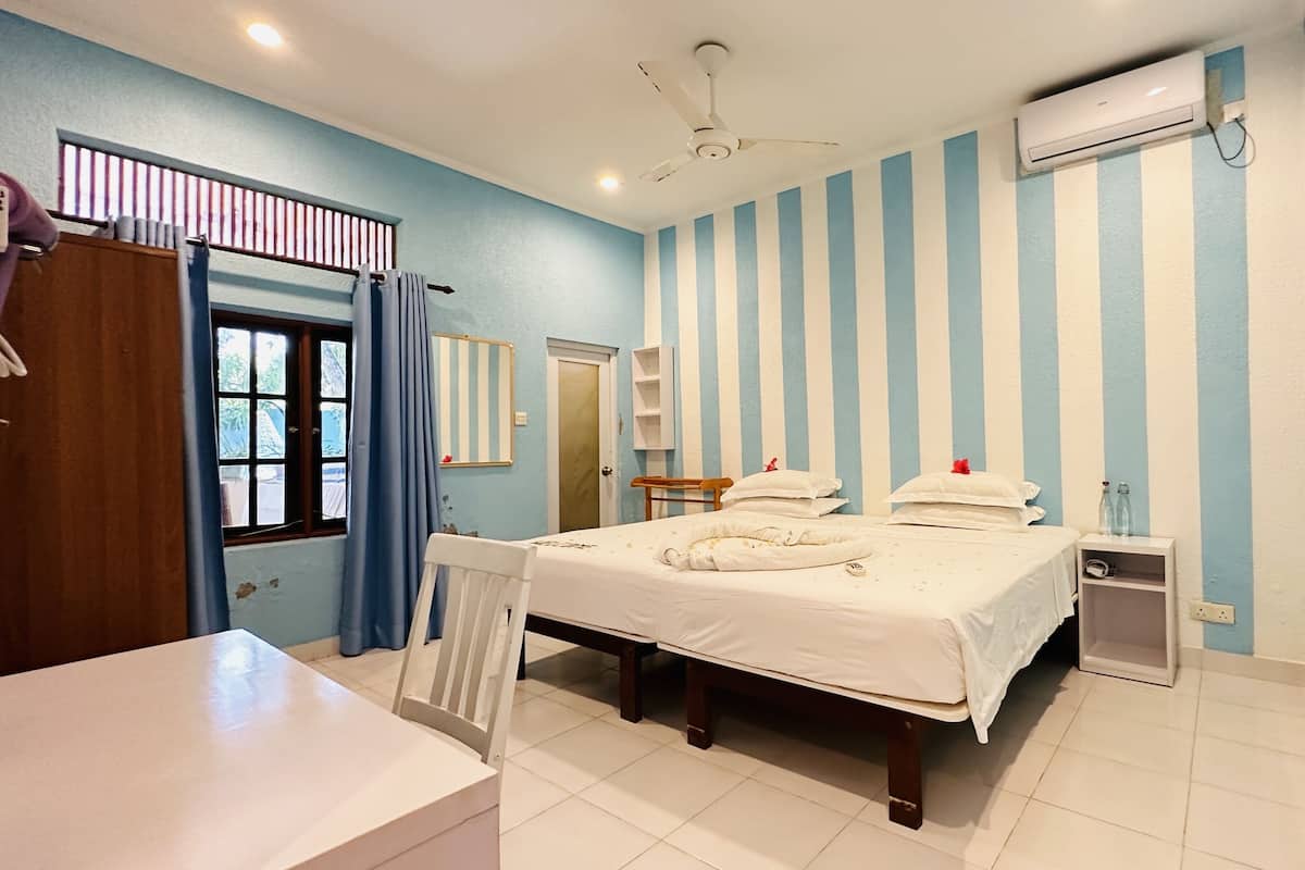 A hotel room with a bed, a desk, a chair, a window with curtains, a ceiling fan, an air conditioning unit, and a small bedside table with bottles.