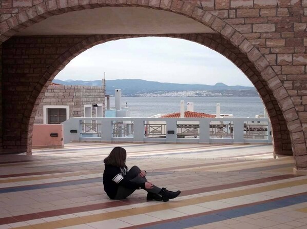 A person sitting under a stone archway overlooking a waterfront with buildings and a view of the sea.