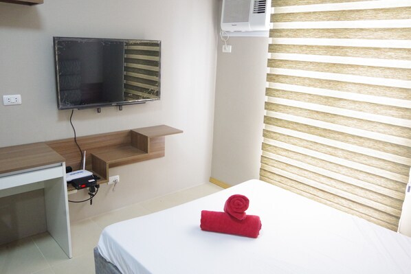 Ein Hotelzimmer mit einem Bett, einem Schreibtisch mit Computer, einem an der Wand befestigten Fernseher und einem Fenster mit Jalousien.