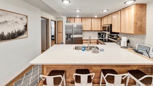 Une cuisine moderne avec des meubles en bois, un grand îlot central doté de tabourets, des appareils en acier inoxydable et des spots encastrés au plafond.