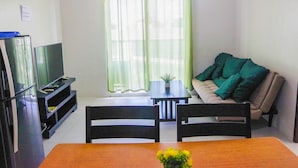 A living room with a TV on a stand, a dining table with chairs, a sofa with a green blanket, and a window with sheer curtains. - Spianada Residences (Cebu City)