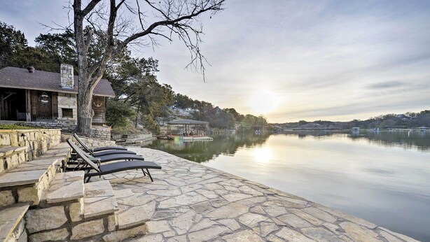 Una terraza de piedra con sillones de descanso con vista a un lago tranquilo, un pabellón y un edificio de piedra con chimenea.