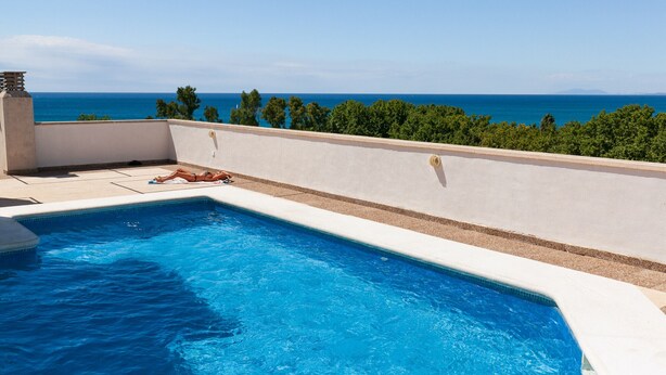 Ein Pool mit Meerblick, eine Person entspannt am Poolufer, und ein klarer Himmel.