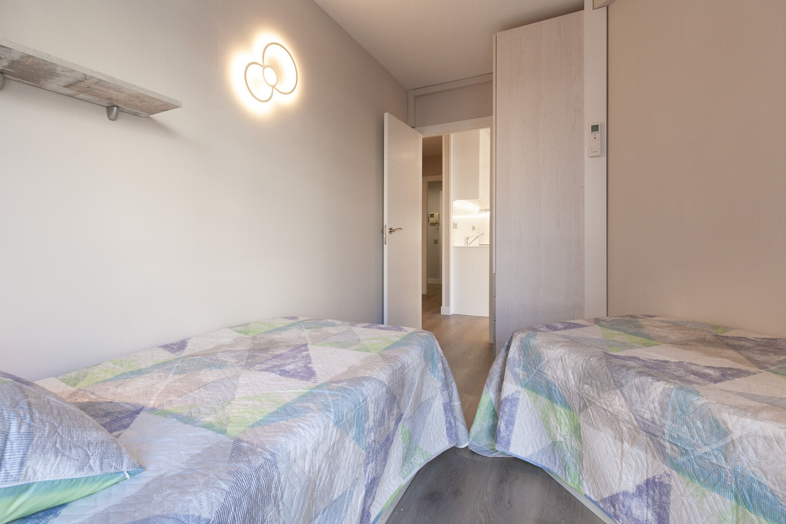 A bedroom with two beds, a bedside table, a wall-mounted light, and a door leading to another room.