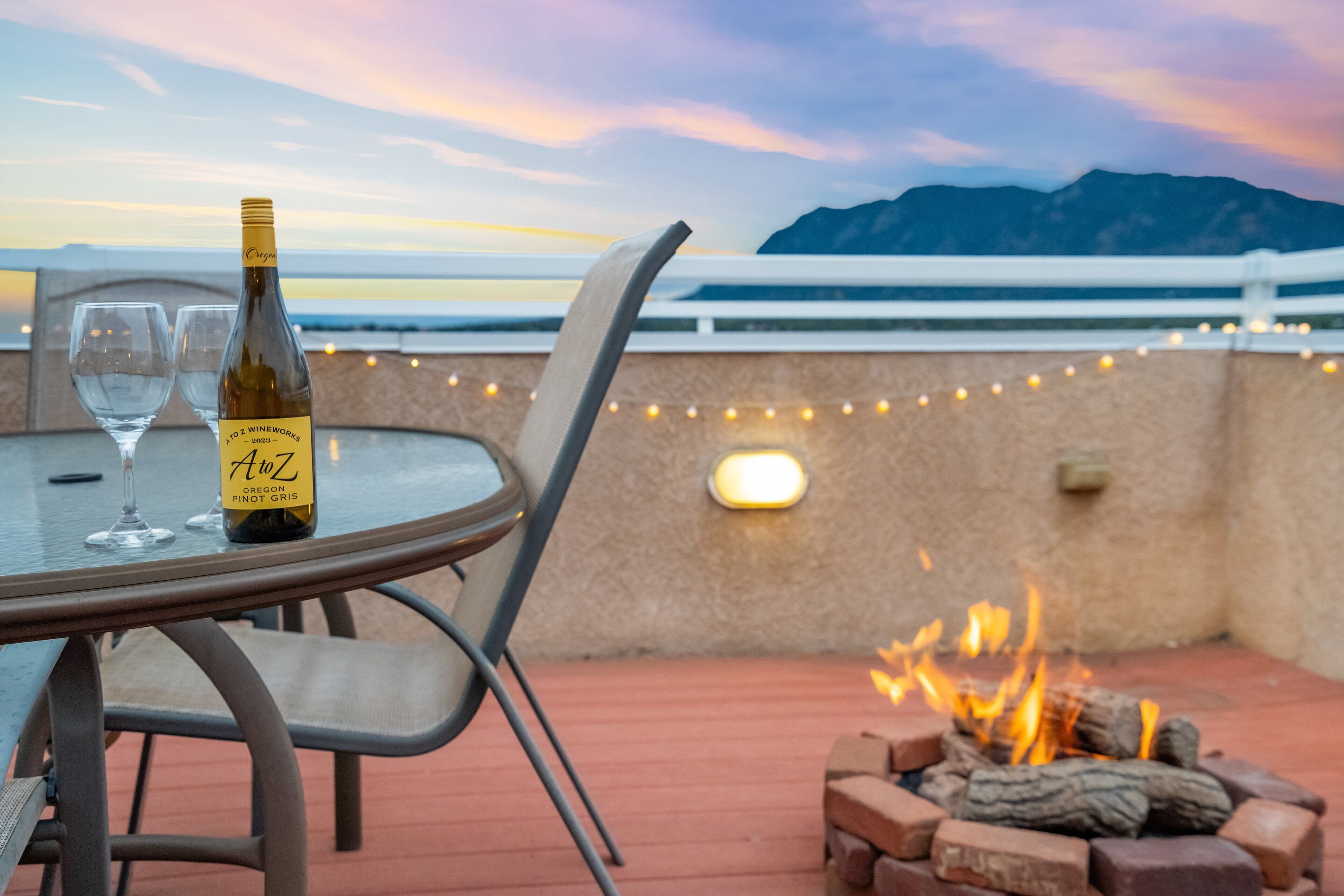 A table with a bottle of wine and glasses, a fire pit, and a view of the ocean at sunset.