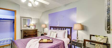 Une chambre à coucher avec une couvre-lit violette, un ventilateur de plafond, une table de chevet avec une lampe et une commode.
