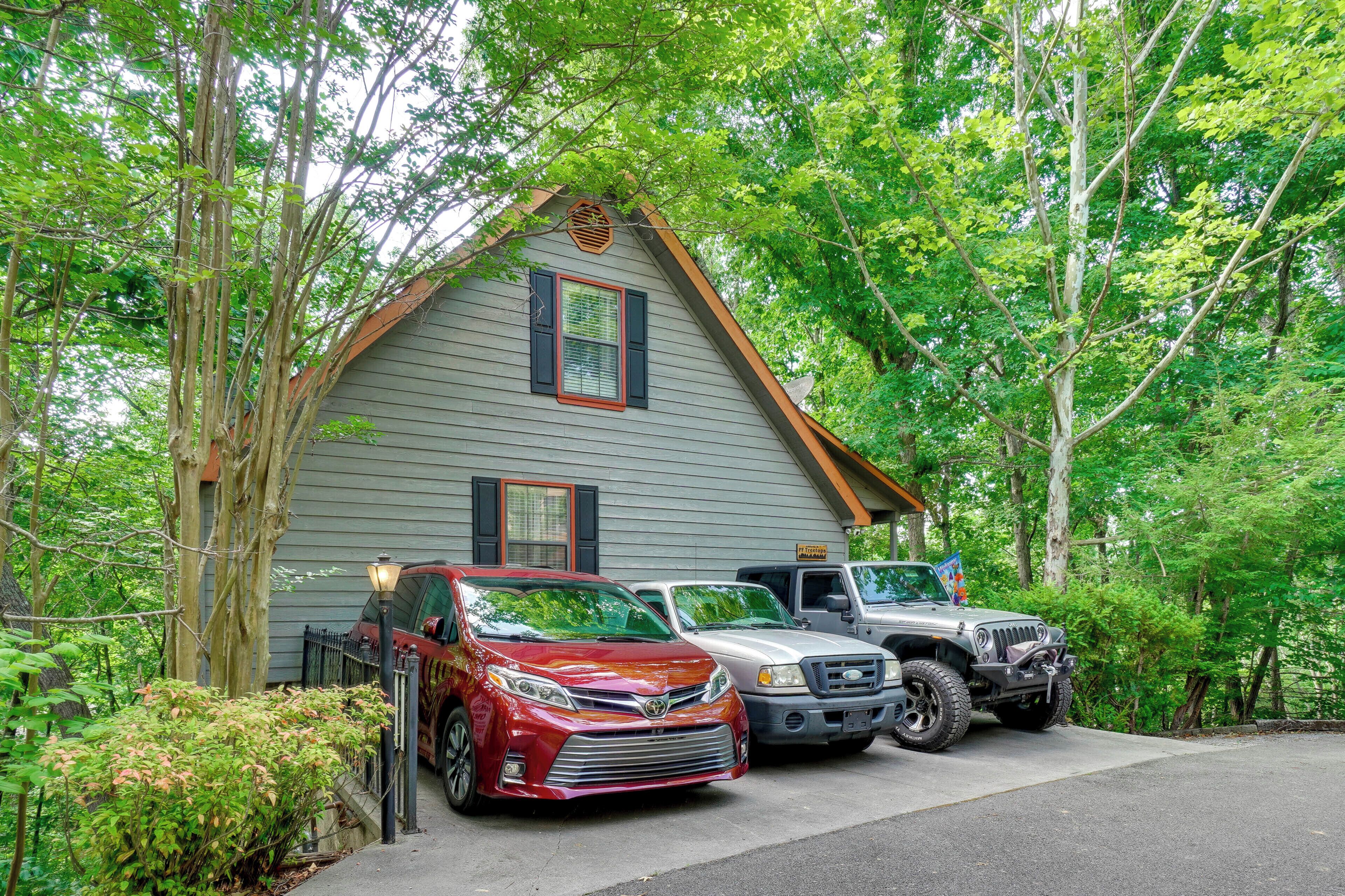 A house with a sloped roof and a garage. There are three vehicles parked in front: a red car, a white car, and a truck.