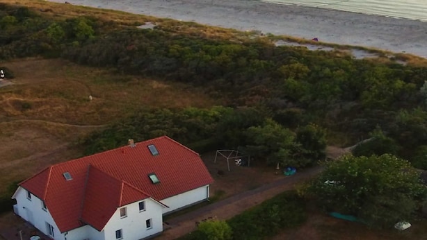 Une maison blanche avec un toit rouge, entourée d’une végétation luxuriante et donnant sur une plage.