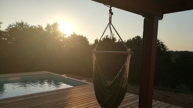 Une terrasse en bois avec un hamac suspendu, une piscine et une vue dégagée sur le coucher du soleil.