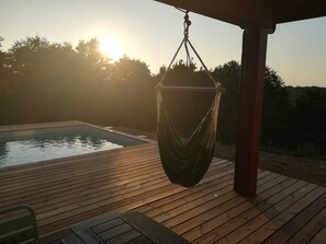 Una terraza de madera con una hamaca colgante, una piscina y una vista despejada del atardecer.
