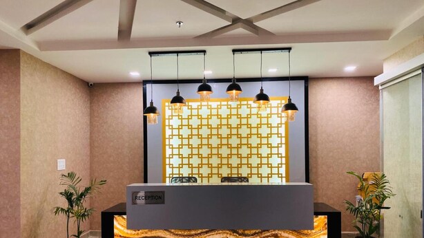 Un comptoir d'accueil au design évoquant le marbre, une enseigne indiquant « RÉCEPTION », et un mur décoré derrière. Trois lampes suspendues sont fixées au-dessus du comptoir, et deux plantes en pot sont placées de chaque côté.