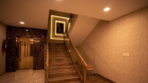 Un escalier moderne avec des marches en bois et des rampes en verre, menant à un ascenseur dont le mur est en marbre et sur le côté gauche se trouve un extincteur.