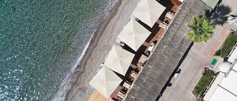 Vista aérea de un hotel frente a la playa con una hilera larga de habitaciones, una terraza de madera y una palmera.