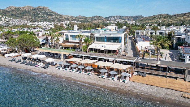 Un hotel en la costa con ubicación frente a la playa, que incluye tumbonas y sombrillas, y vistas al mar y colinas al fondo.