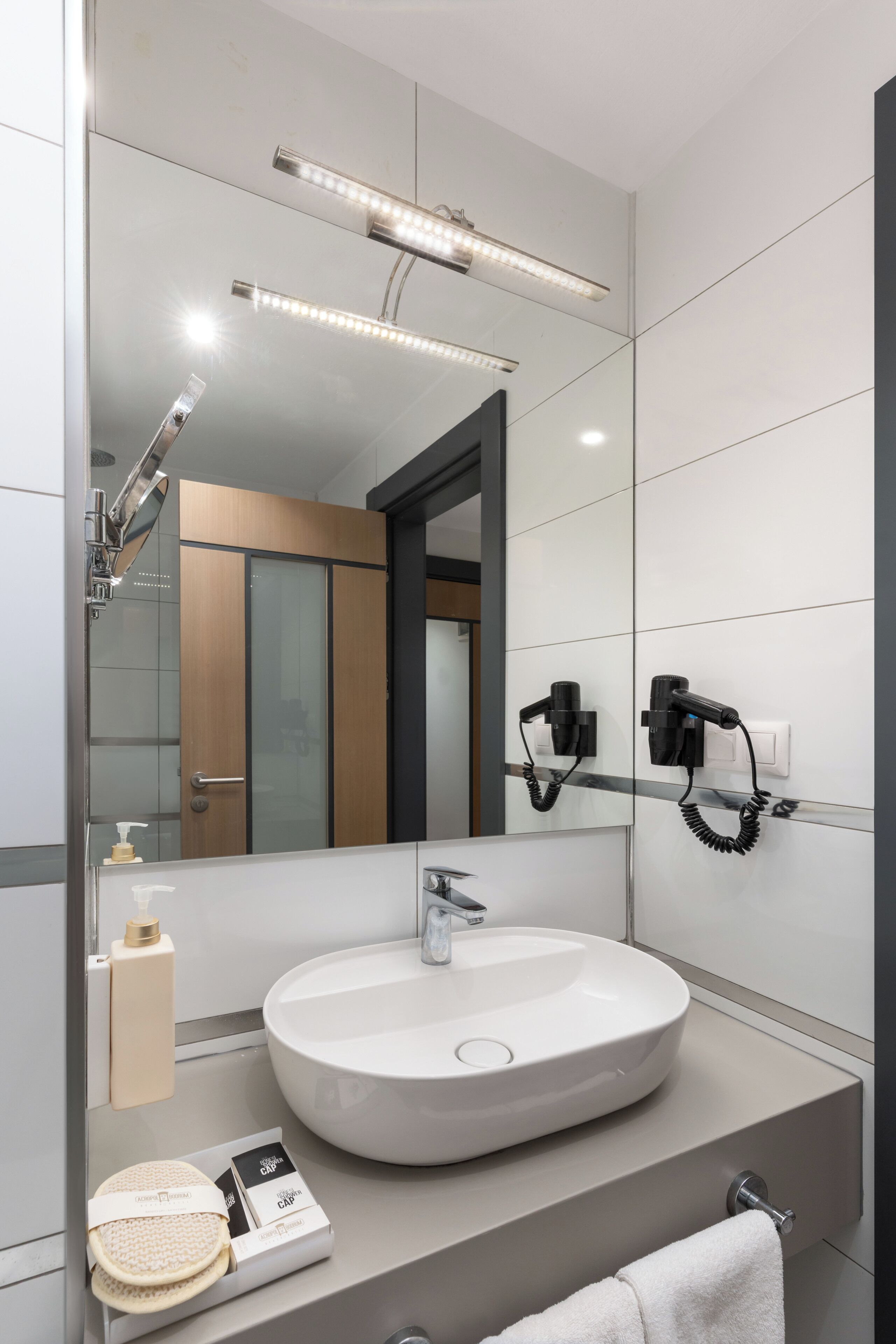A modern bathroom with a large mirror, a round sink, a handheld showerhead, and a hairdryer mounted on the wall.