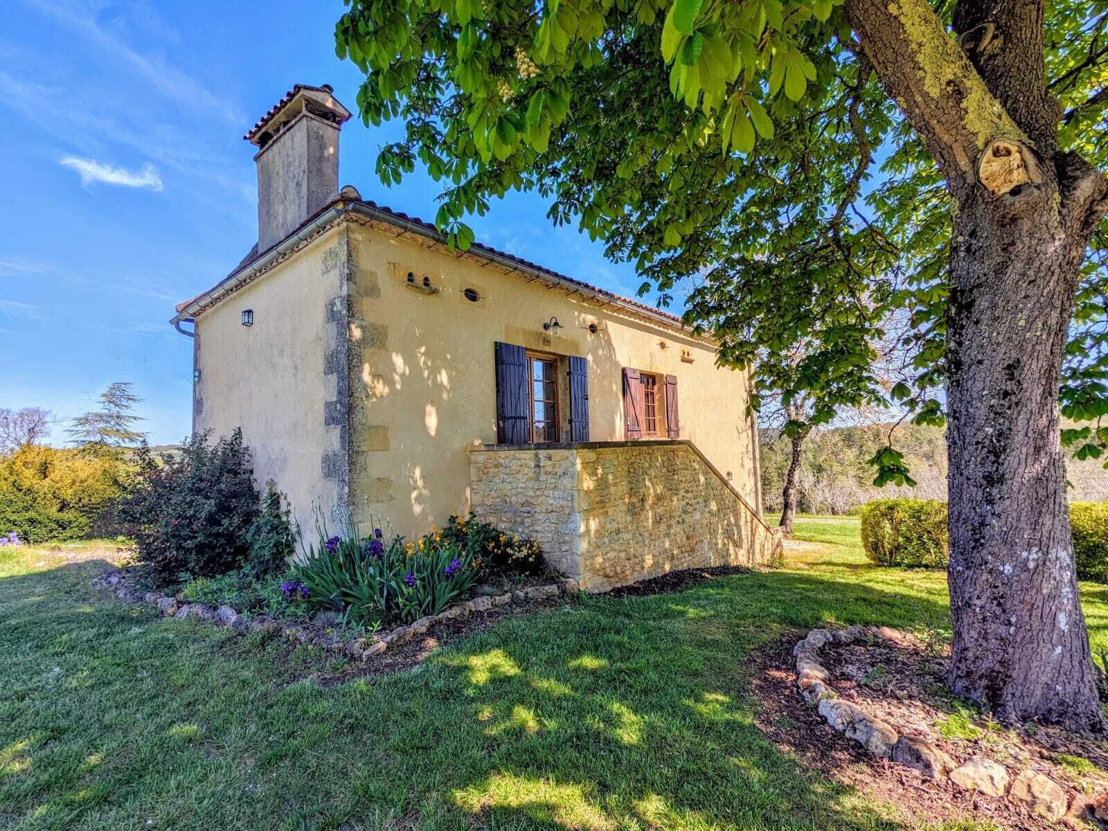 A rustic stone house with a chimney, wooden shutters, and a small garden with flowers and shrubs.
