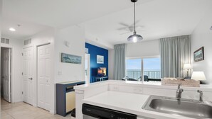 Cocina moderna con encimeras blancas, lavavajillas y pileta. Una pared de acento azul con un cuadro y una ventana con cortinas.
