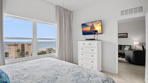 Habitación de hotel con una cama grande, televisor montado en la pared, vista a la playa y un área de estar independiente con sofá y lámpara.