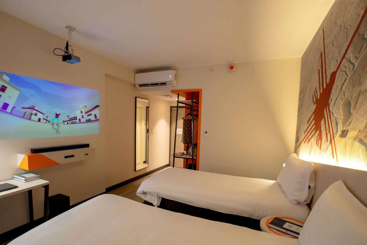 Une chambre d’hôtel avec deux lits, un bureau avec une lampe, un écran de projection, un appareil de climatisation et un tableau au mur.