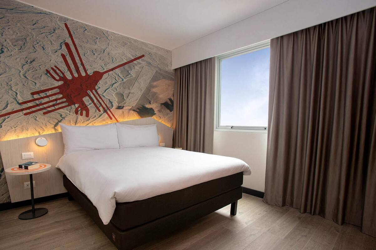 Une chambre d’hôtel avec un grand lit, une table de chevet, une lampe fixée au mur et une fenêtre avec des rideaux.