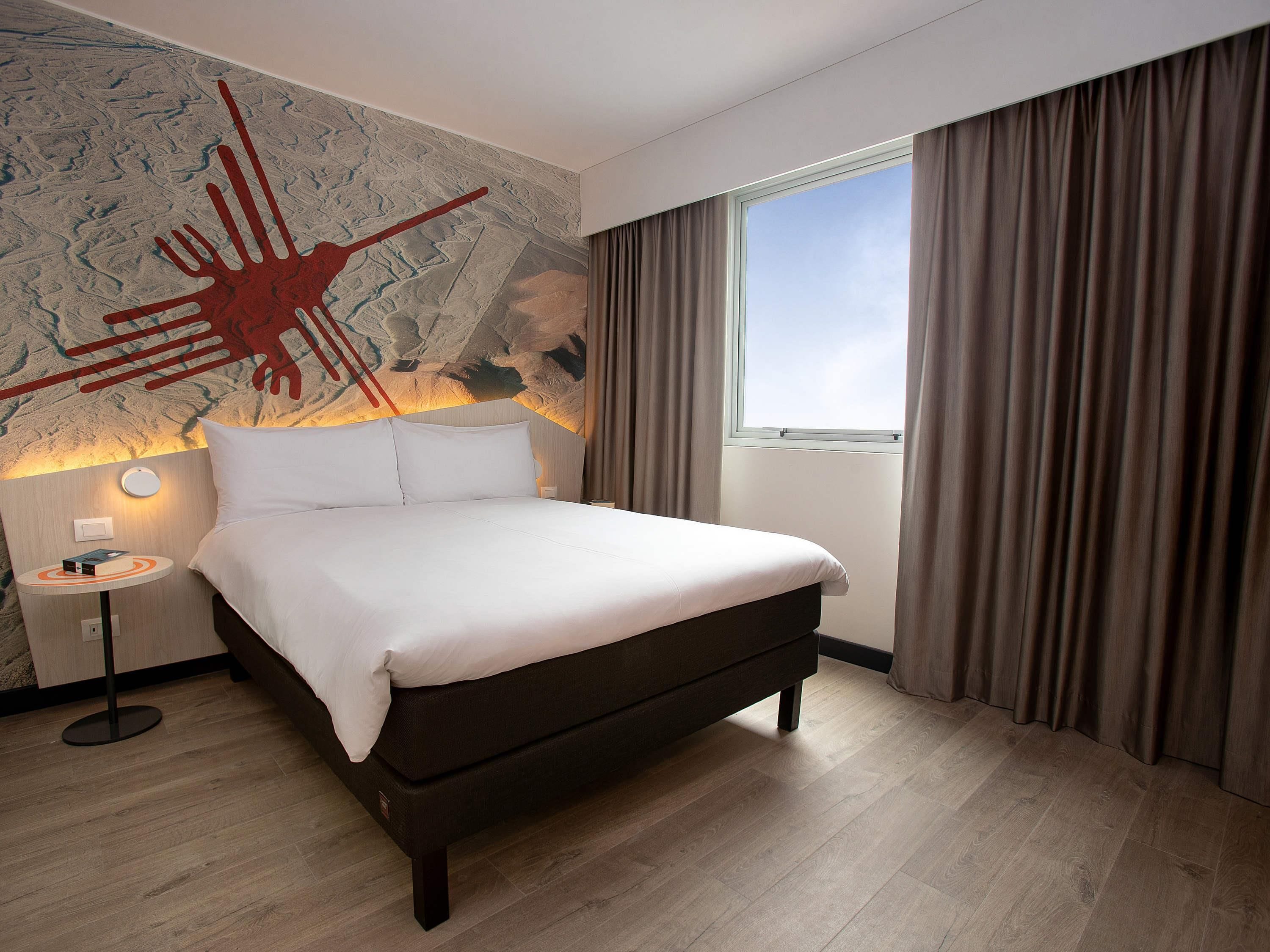 Quarto de hotel com uma cama grande, uma mesa de cabeceira, uma luminária na parede e uma janela com cortinas.