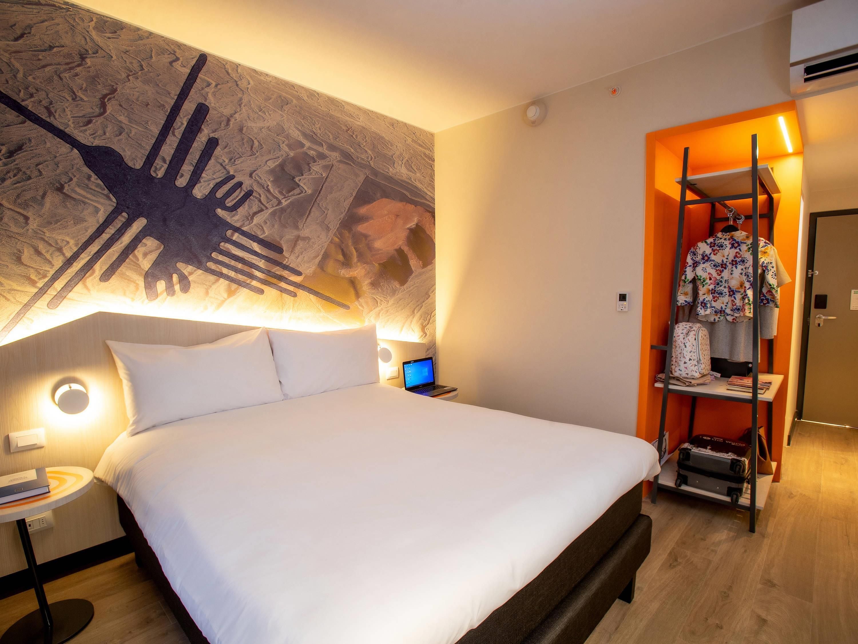 A hotel room with a large bed, a wall-mounted artwork, a small desk with a laptop, and a wardrobe with a hanging rack.