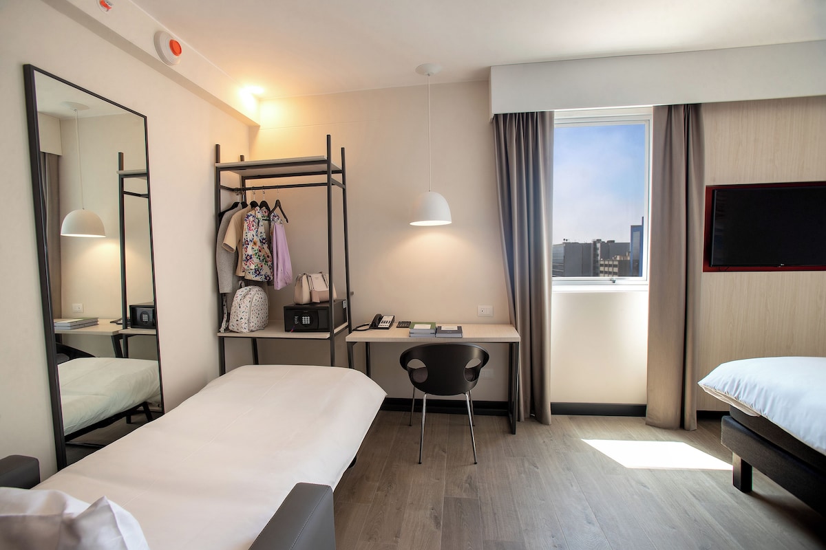 Une chambre d’hôtel équipée d’un lit, d’un bureau, d’une chaise, d’une armoire, d’un miroir et d’une fenêtre donnant sur la ville.