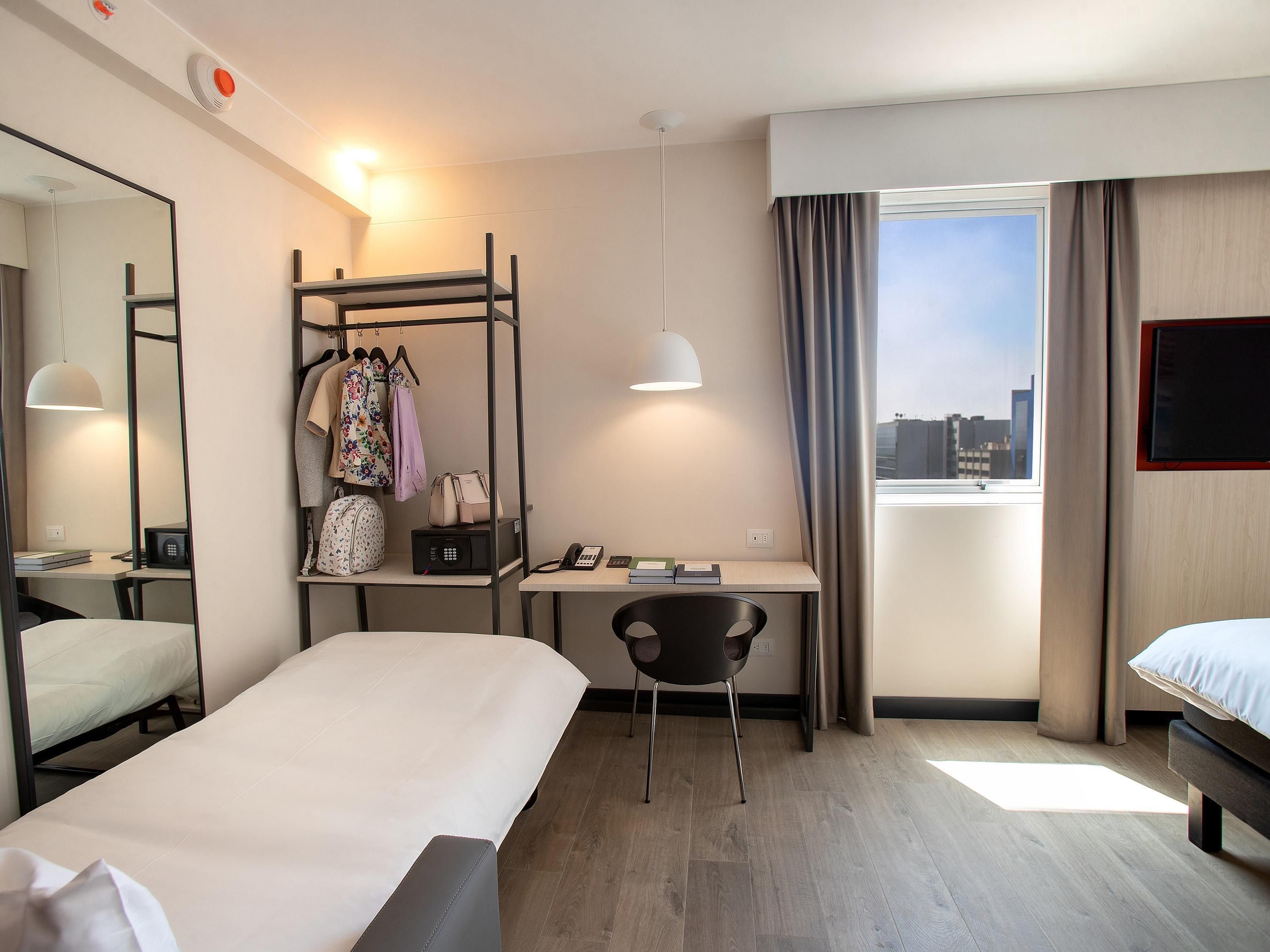 Quarto de hotel com cama, escrivaninha, cadeira, guarda-roupa e janela com vista para a cidade.