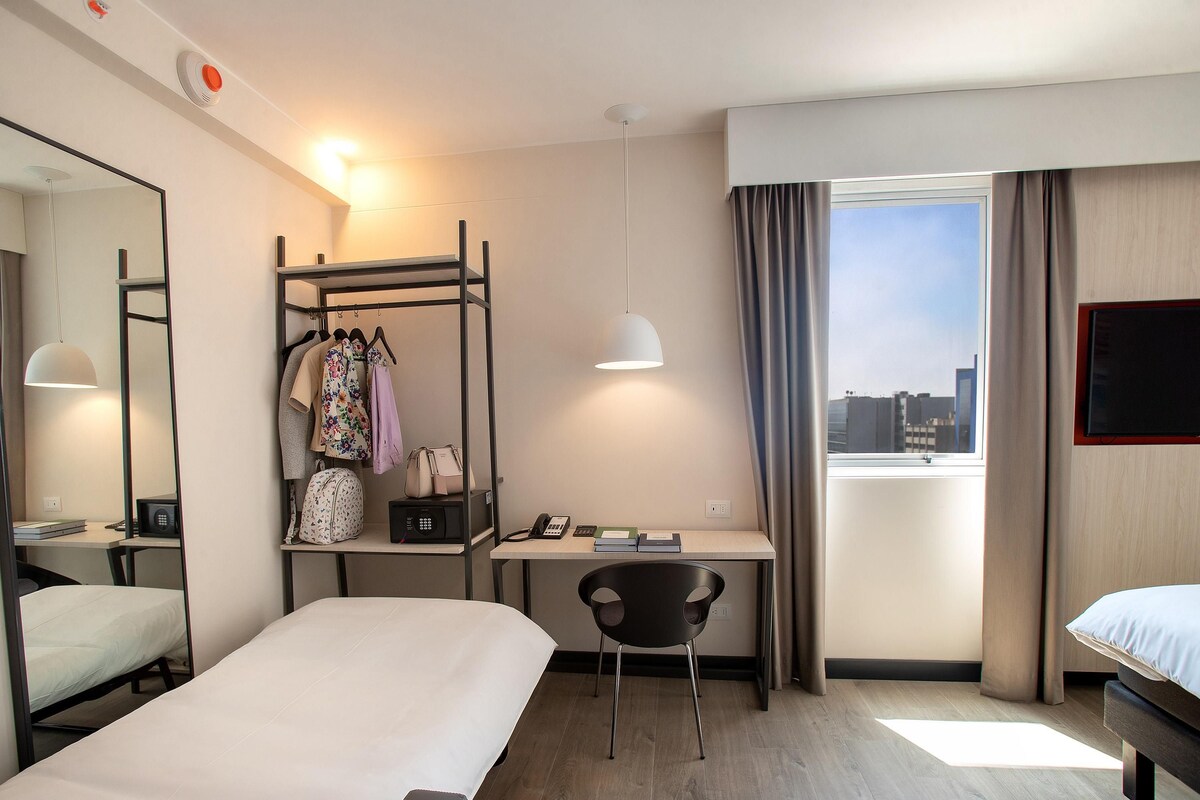 Een hotelkamer met een bed, een bureau, een stoel, een kledingkast en een raam met uitzicht over de stad.