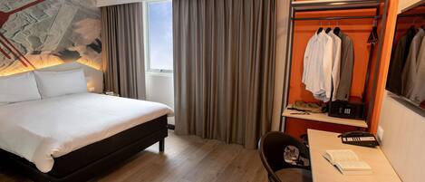 A hotel room with a bed, a desk with a chair, a coat rack with clothes, and a window with curtains.