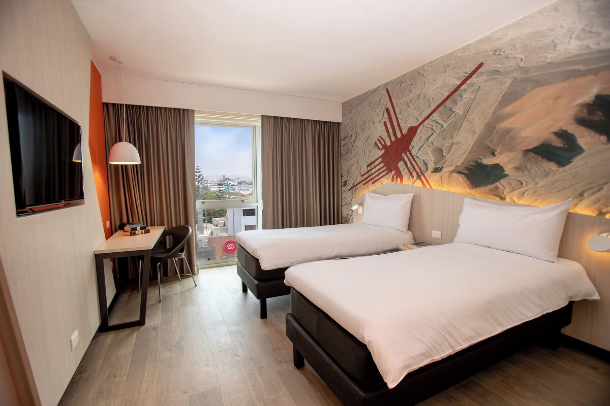 Une chambre d’hôtel avec deux lits, un bureau et une grande fenêtre avec des rideaux.