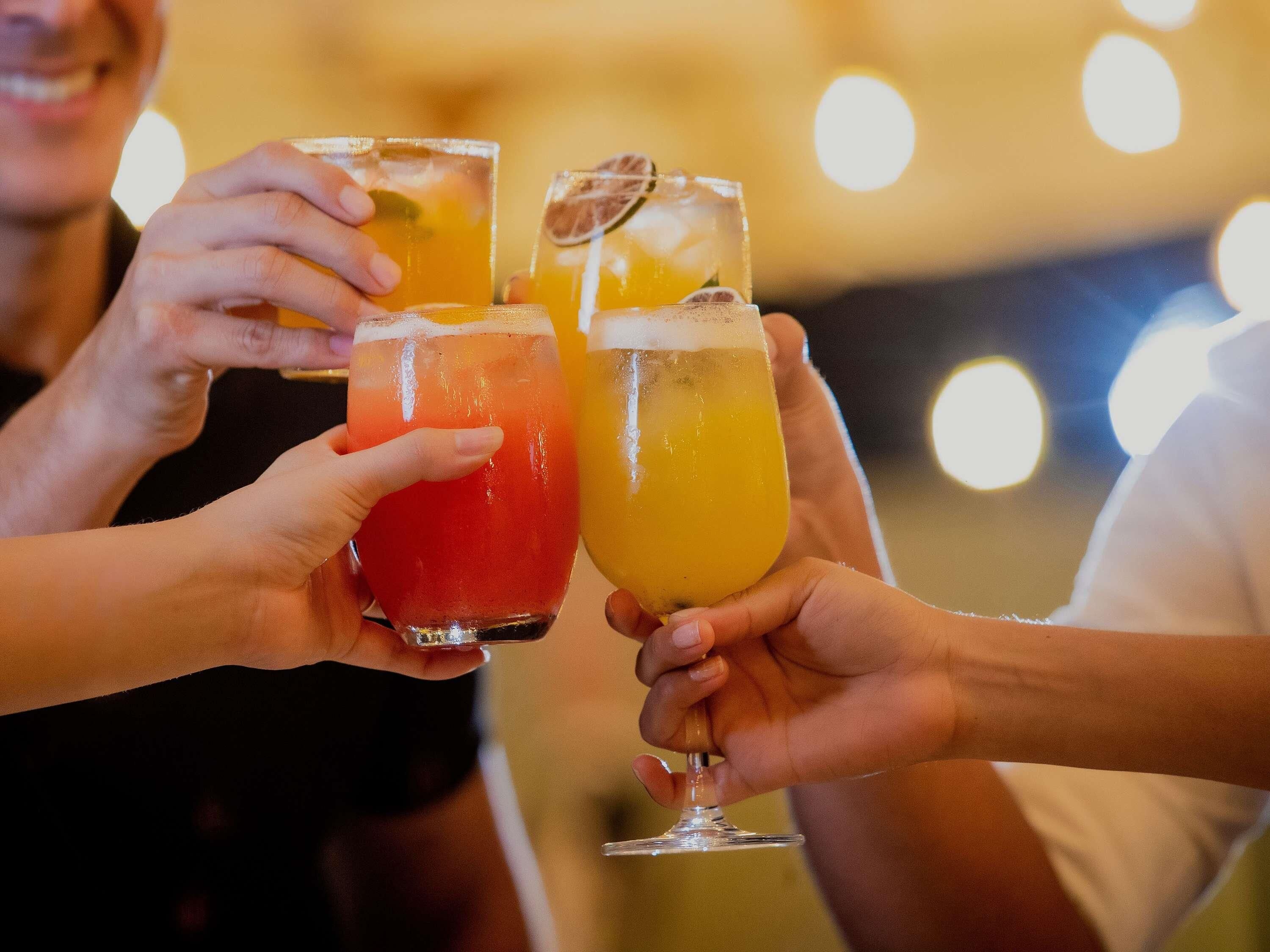 Three people holding glasses of different colored drinks, likely cocktails, in a dimly lit setting with blurred lights in the background.